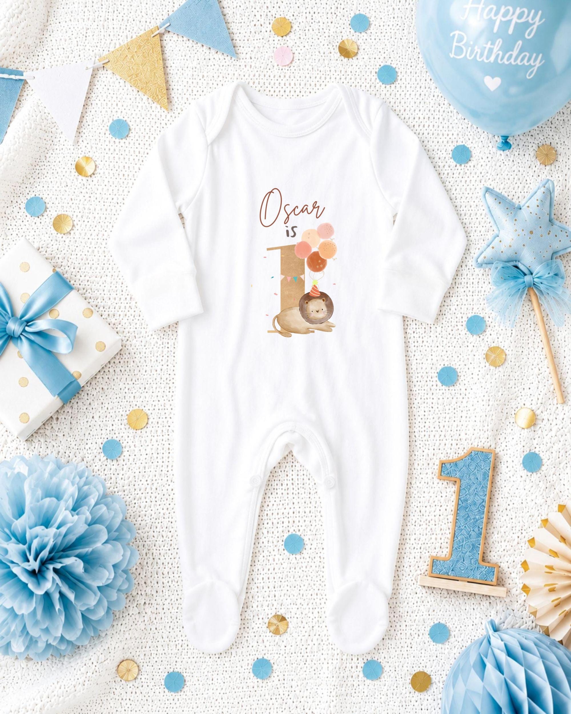 A white onesie with the name "Oscar" printed on it, surrounded by various baby-themed items such as a blue balloon, a blue and gold star, and blue and gold confetti.