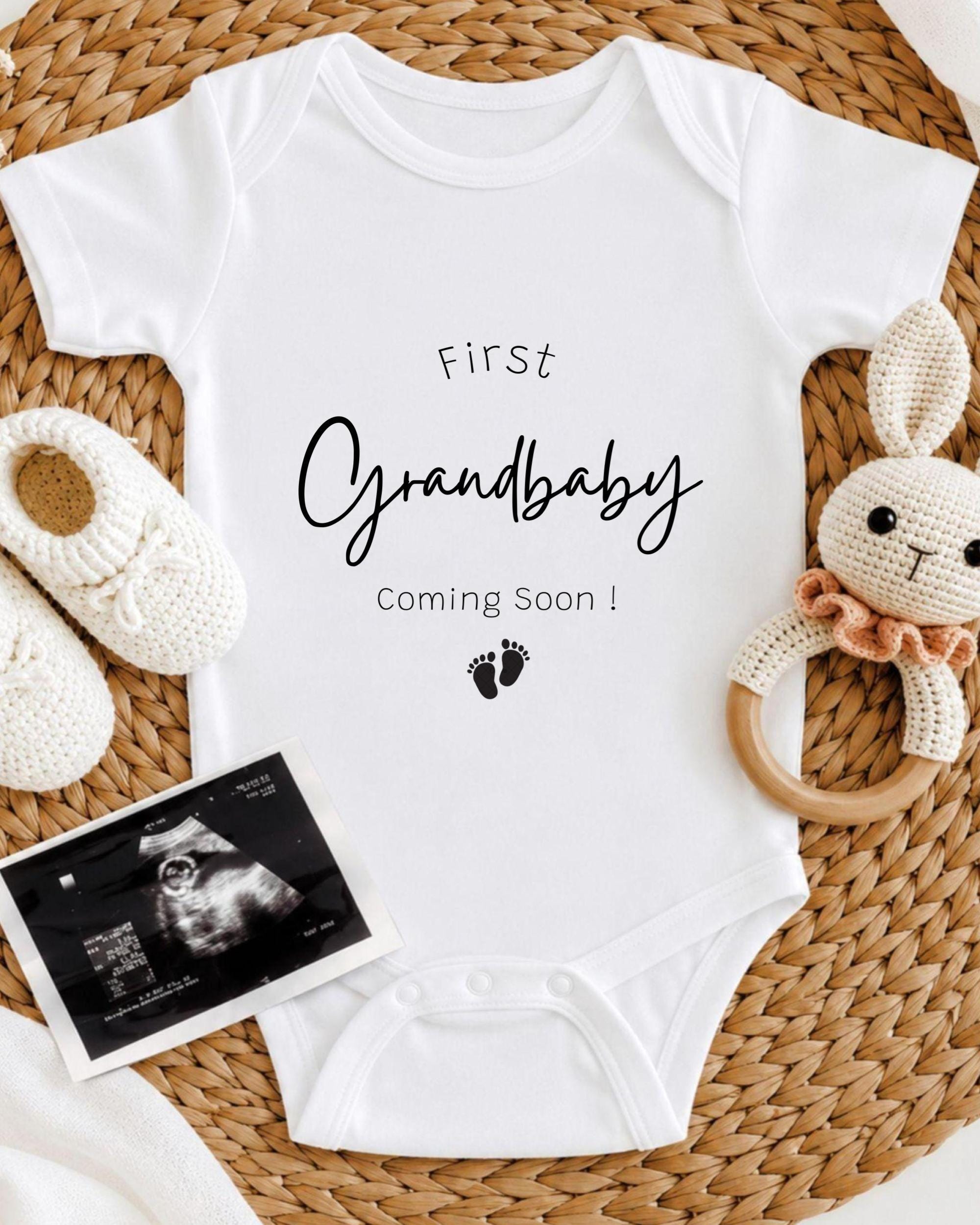 A white onesie with the text "First Grandbaby Coming Soon!" printed on it, along with a crocheted bunny toy and a black and white photograph of an infant's belly.