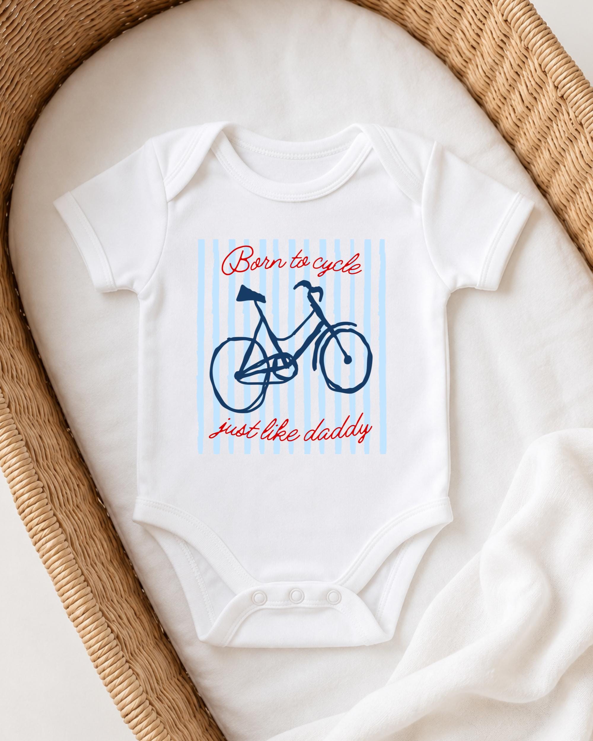 A white baby onesie with a bicycle graphic and the text "Born to cycle just like daddy" printed on it, placed in a woven basket.