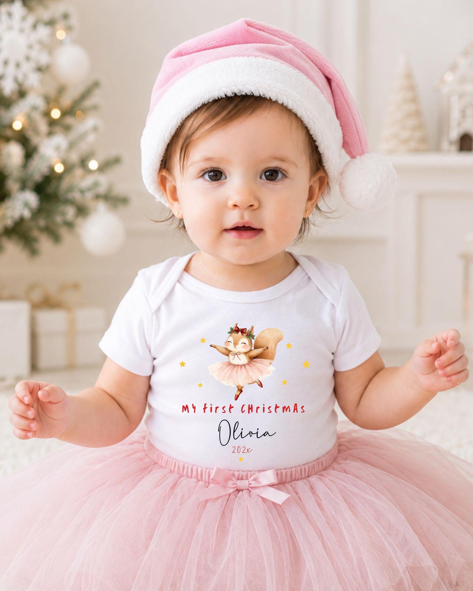 A young girl wearing a pink Santa hat and a white t-shirt with a cartoon character on it, posing in front of a Christmas tree.