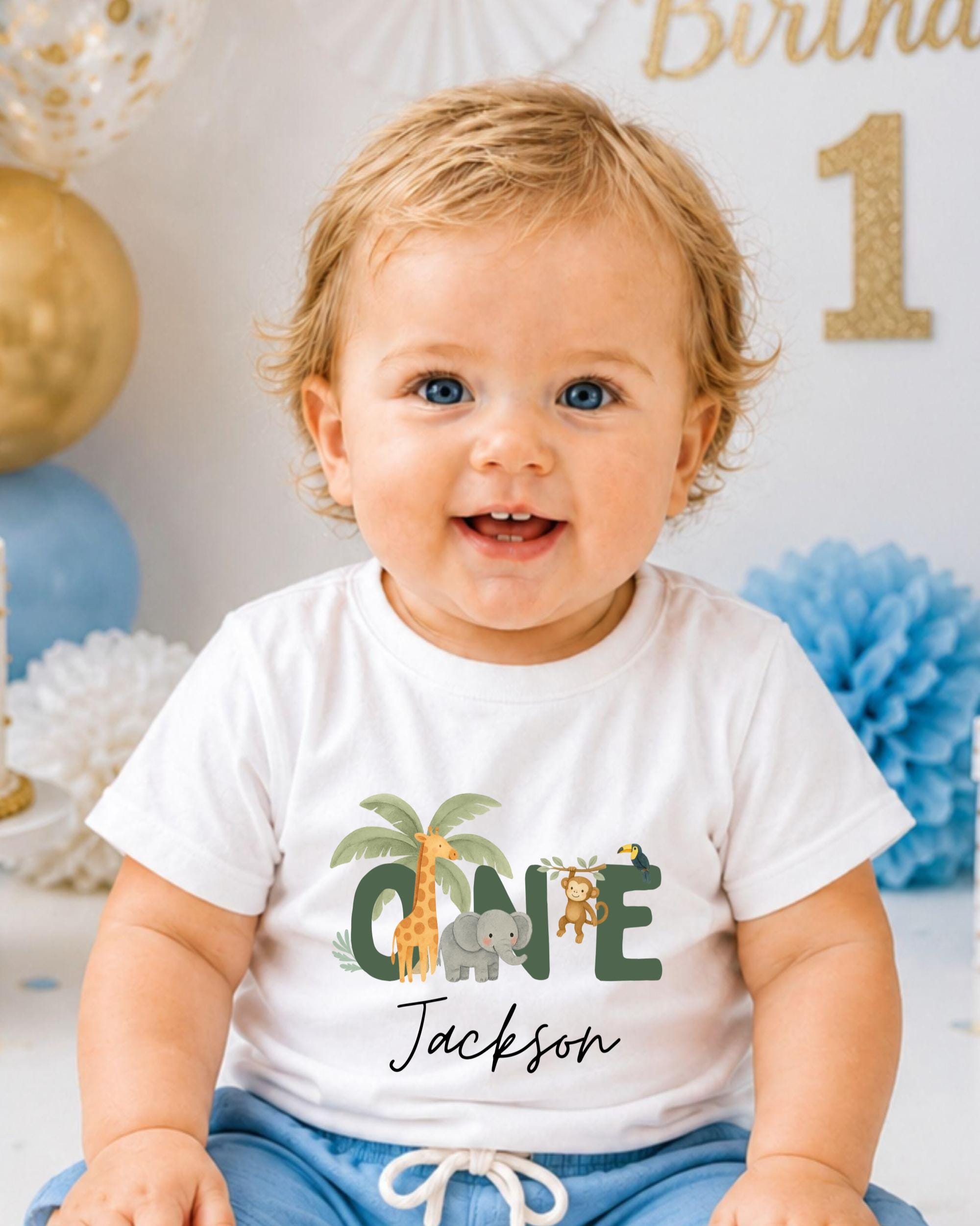 A smiling baby boy wearing a white t-shirt with a jungle-themed design and the name "Jackson" printed on it.