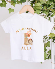 A white t-shirt with a printed design on it, hanging on a wooden hanger. The design features a cartoon image of a lion and the text "My first birthday Alex".