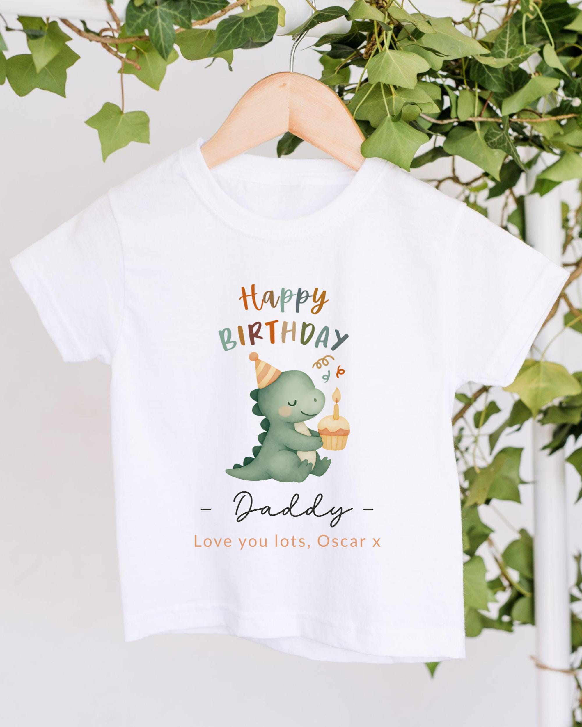 A white t-shirt with a graphic design of a dinosaur wearing a birthday hat and holding a cupcake, along with the text "Happy Birthday Daddy - Love you lots, Oscar x".