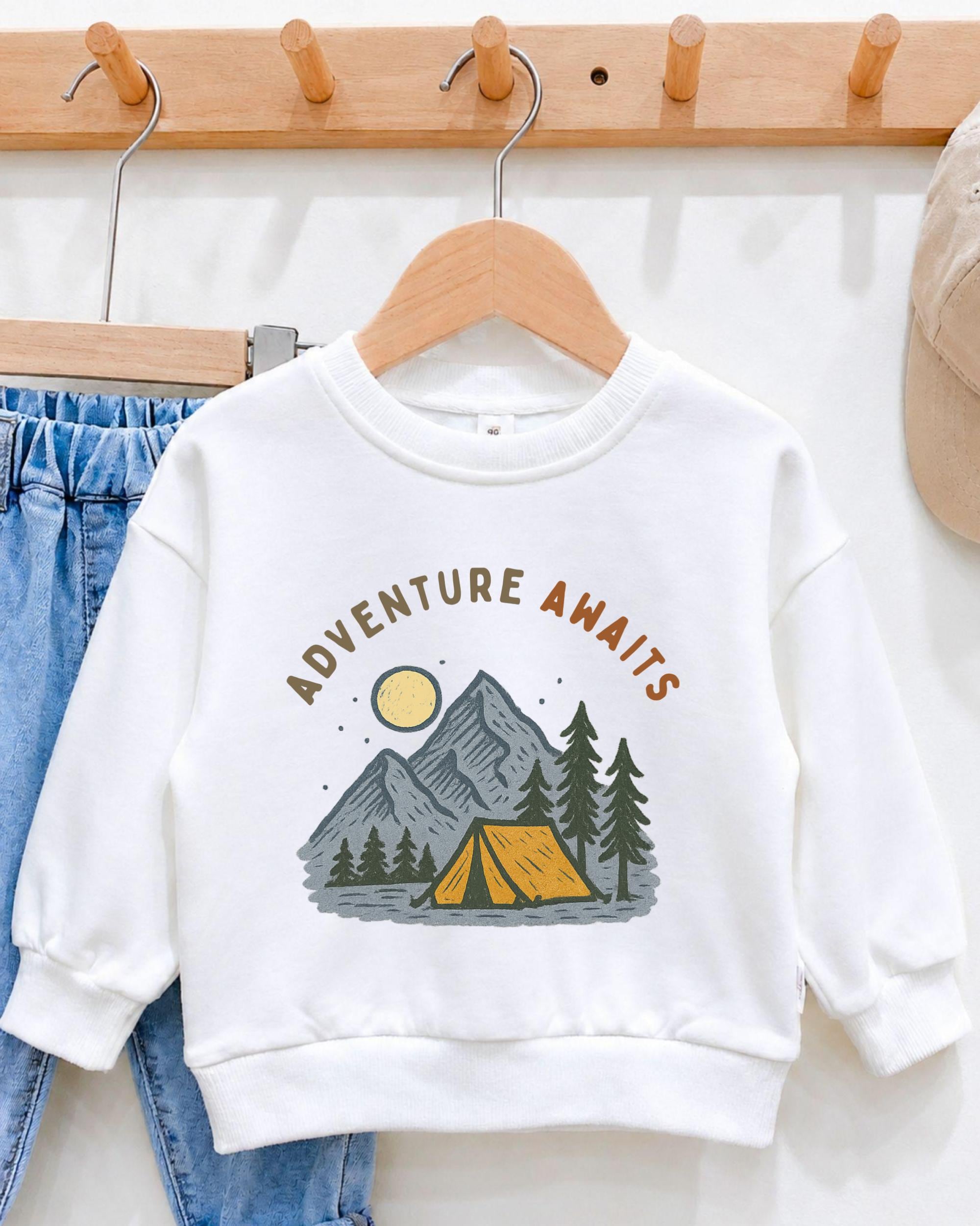 A white sweatshirt with a graphic design of a tent and mountains, along with the text "Adventure Awaits" printed on it, hanging on a wooden hanger.