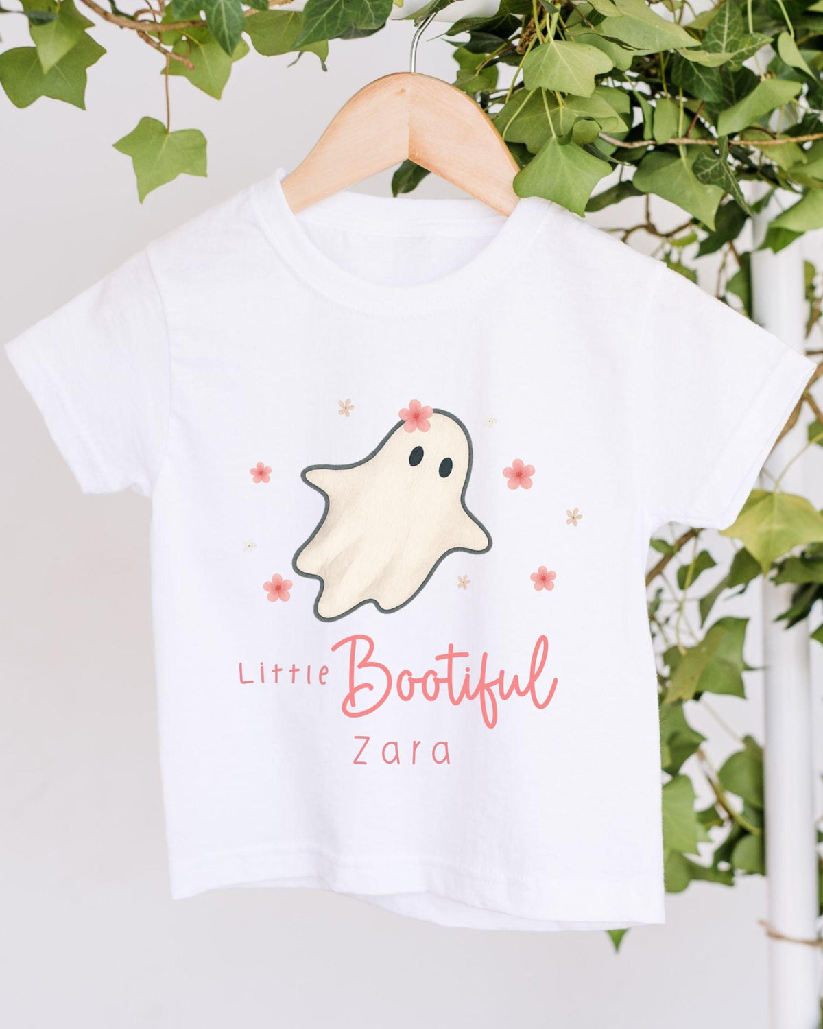 A white t-shirt with a ghost design and the text "Little Bootiful Zara" printed on it, hanging on a wooden hanger.