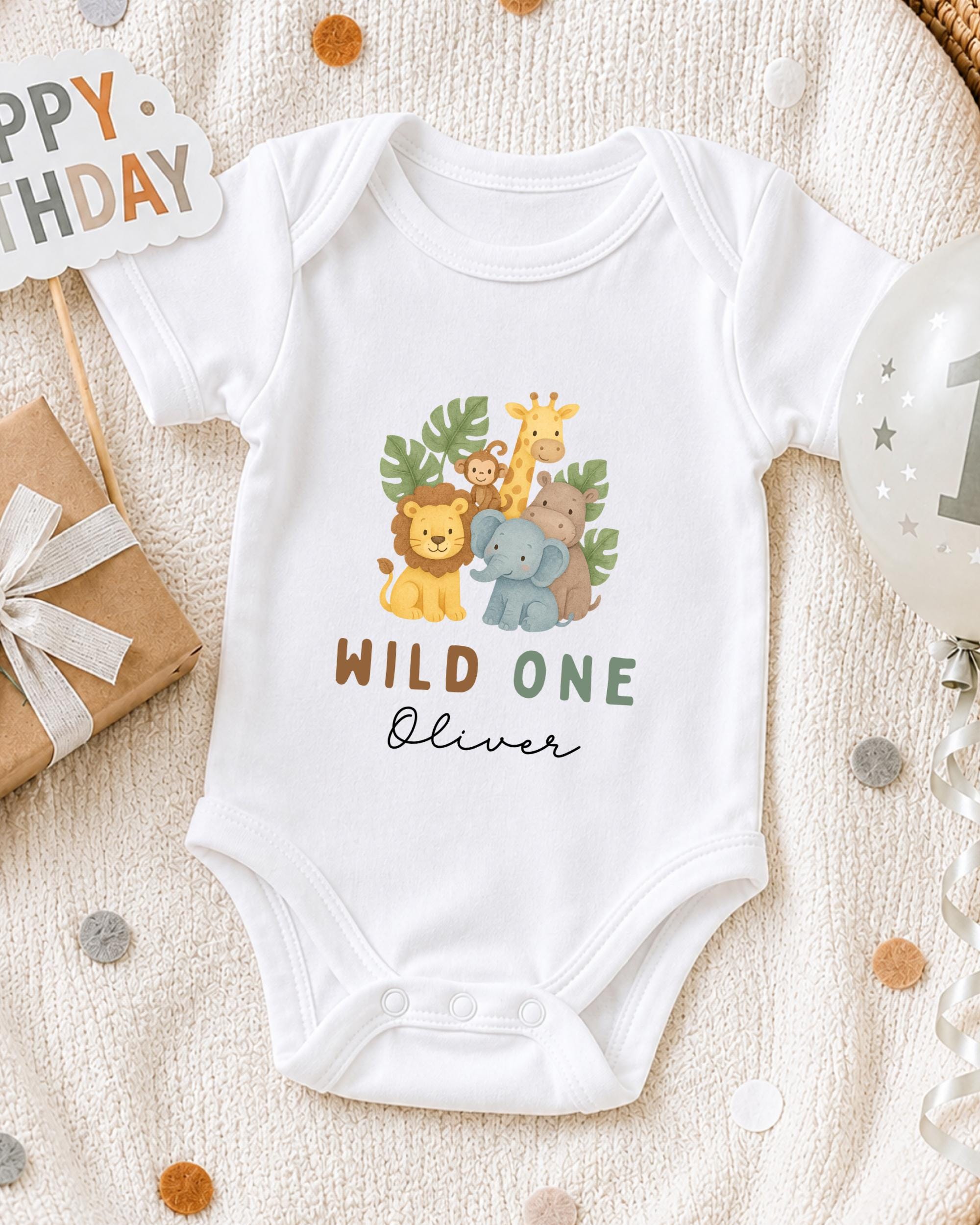 A white onesie with a jungle animal design and the text "Wild One Oliver" printed on it, along with a "Happy Birthday" sign and a balloon.