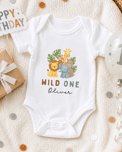 A white bodysuit with a jungle animal design and the text "Wild One Oliver" printed on it, along with a "Happy Birthday" sign and a balloon.