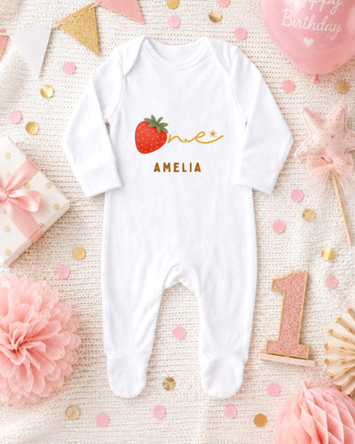 A white bodysuit with a red strawberry and the name "Amalia" printed on it, surrounded by pink and gold decorations, a pink balloon, and a pink flower.