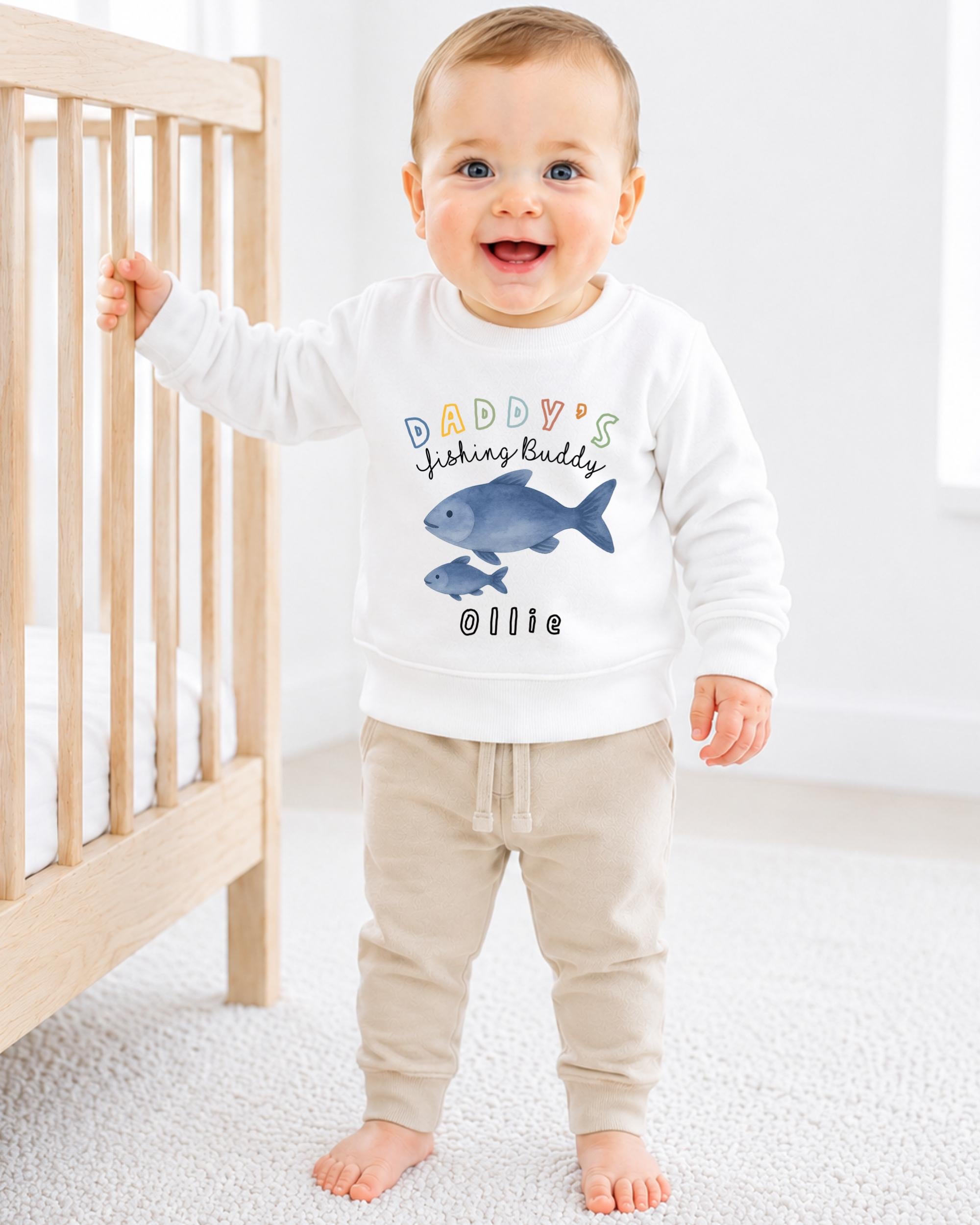 A smiling baby boy wearing a white sweatshirt with a fish design and the text "DADDY'S FISHING BUDDY OLLIE" is standing next to a wooden crib.