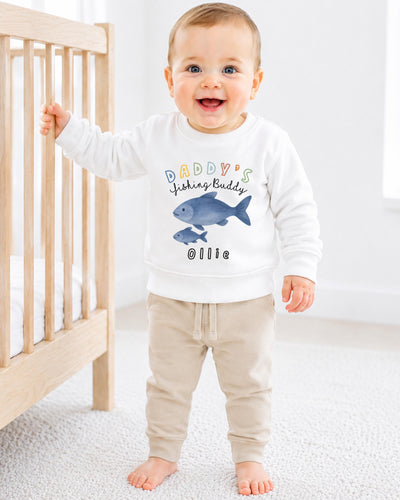 A smiling baby boy wearing a white sweatshirt with a fish design and the text "DADDY'S FISHING BUDDY OLLIE" is standing next to a wooden crib.
