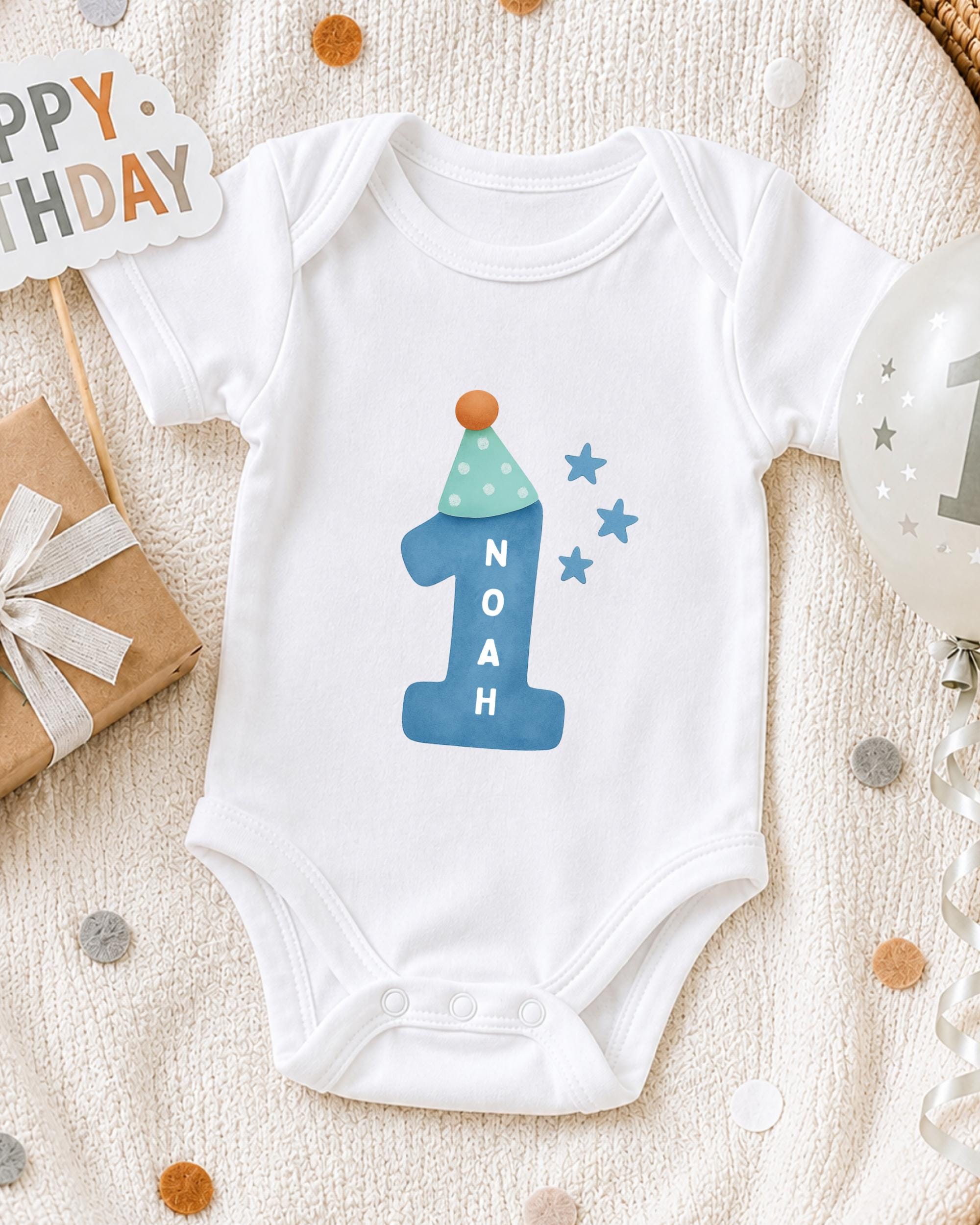 A white onesie with a blue number one and stars on it, along with a "Happy Birthday" sign and a birthday balloon, all placed on a white blanket with confetti.