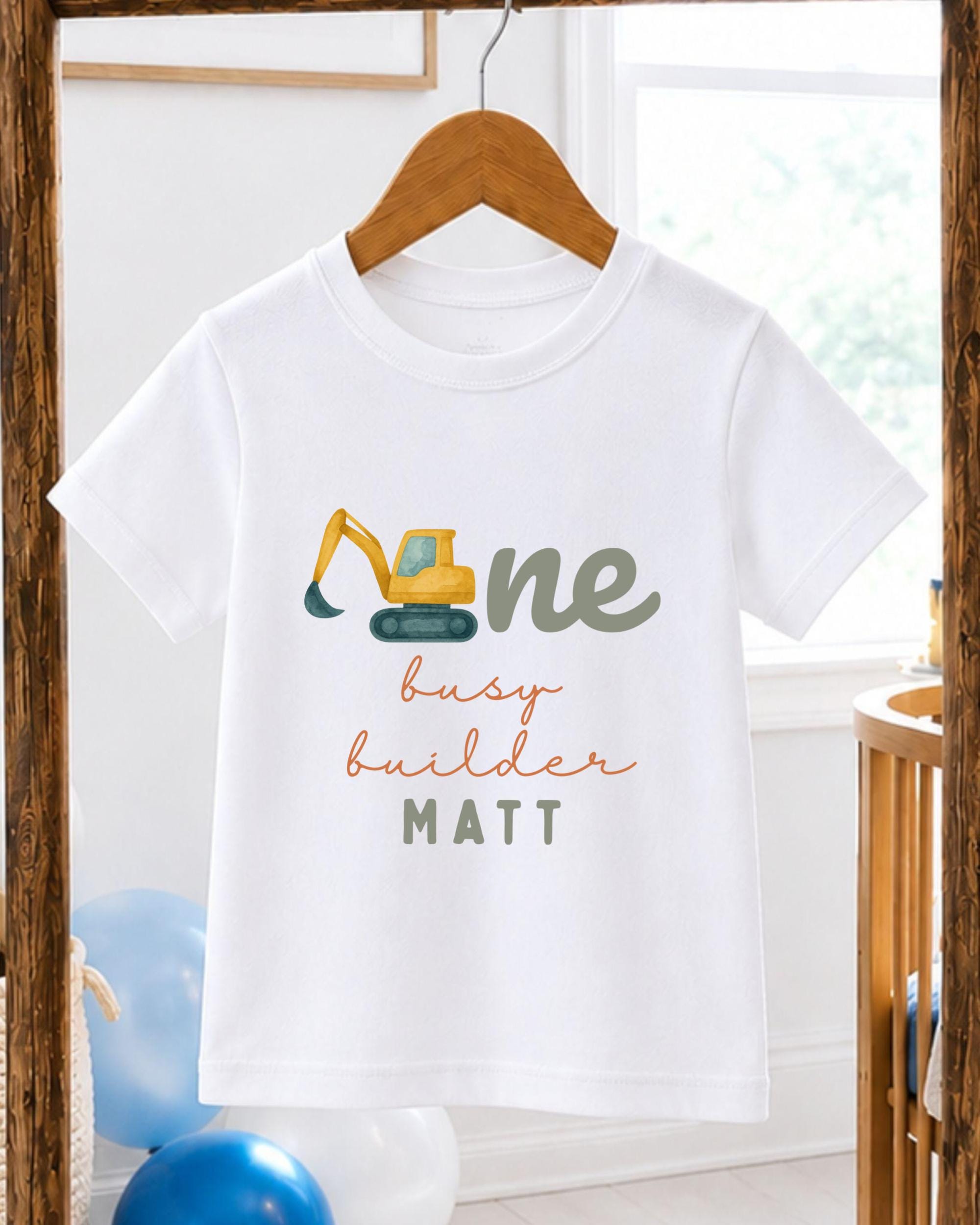 A white t-shirt with a graphic design of a yellow excavator and the text "One busy builder" printed on it.