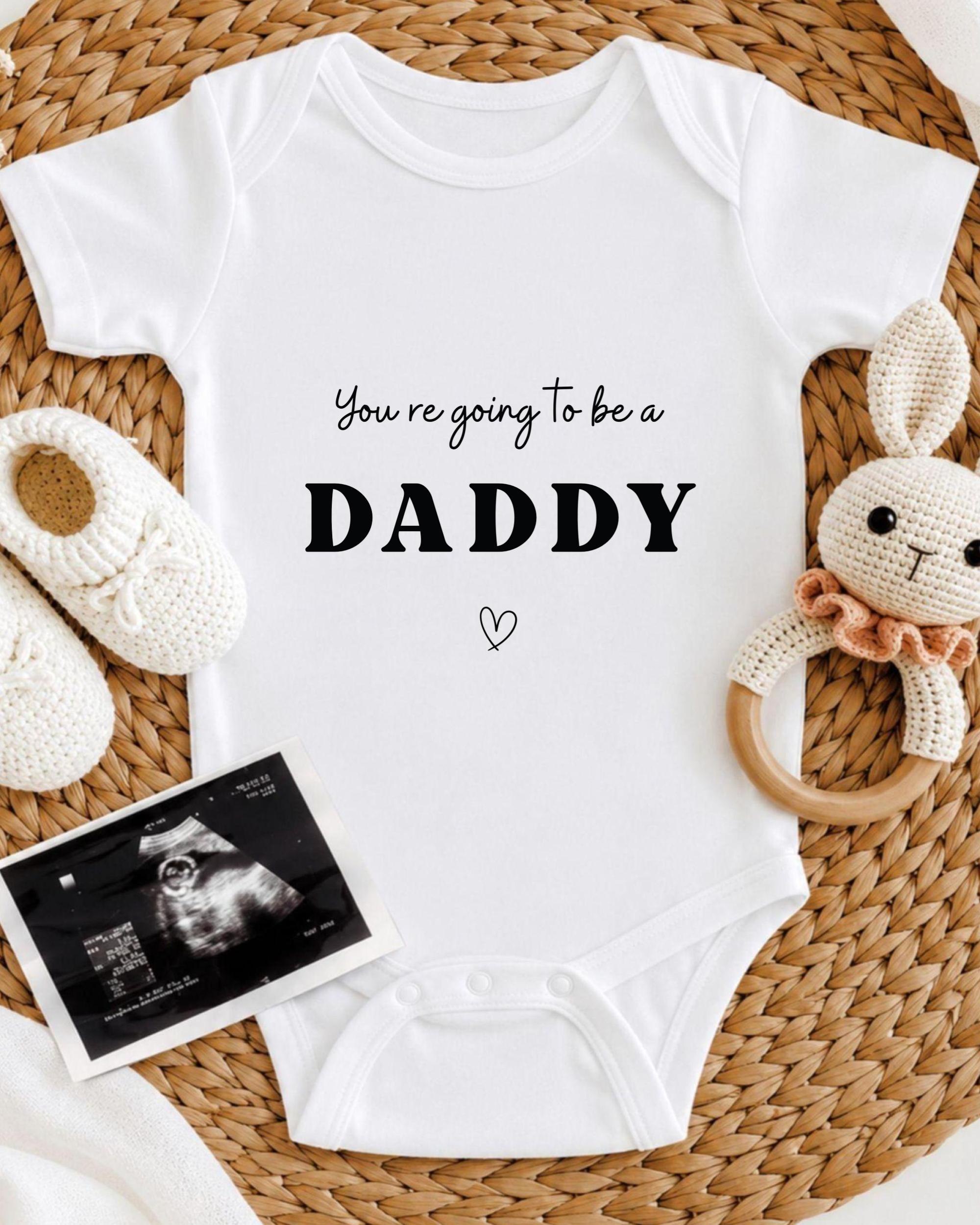 A white baby onesie with the text "You're going to be a DADDY" printed on it, along with a cute stuffed bunny toy and a black and white photograph of an Ultrasound image.