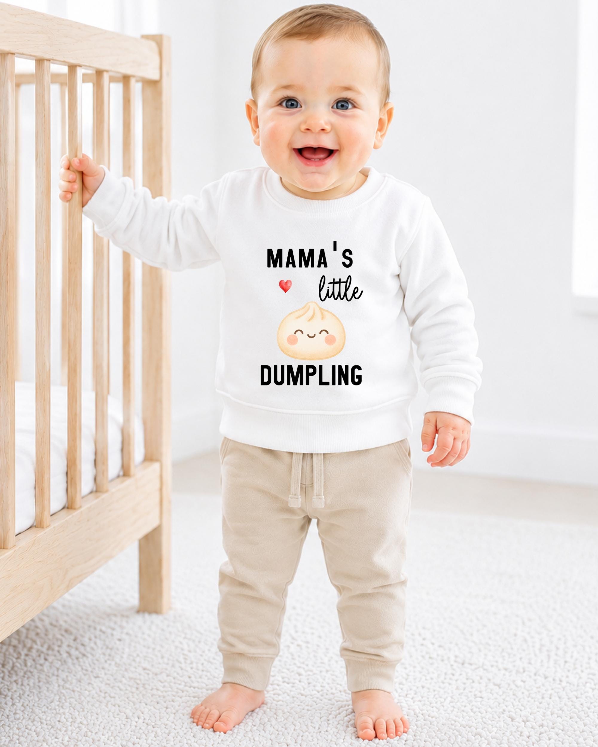 A smiling baby boy wearing a white sweatshirt with a cartoon image of a dumpling and the text "Mama's little DUMPLING" standing in front of a wooden crib.