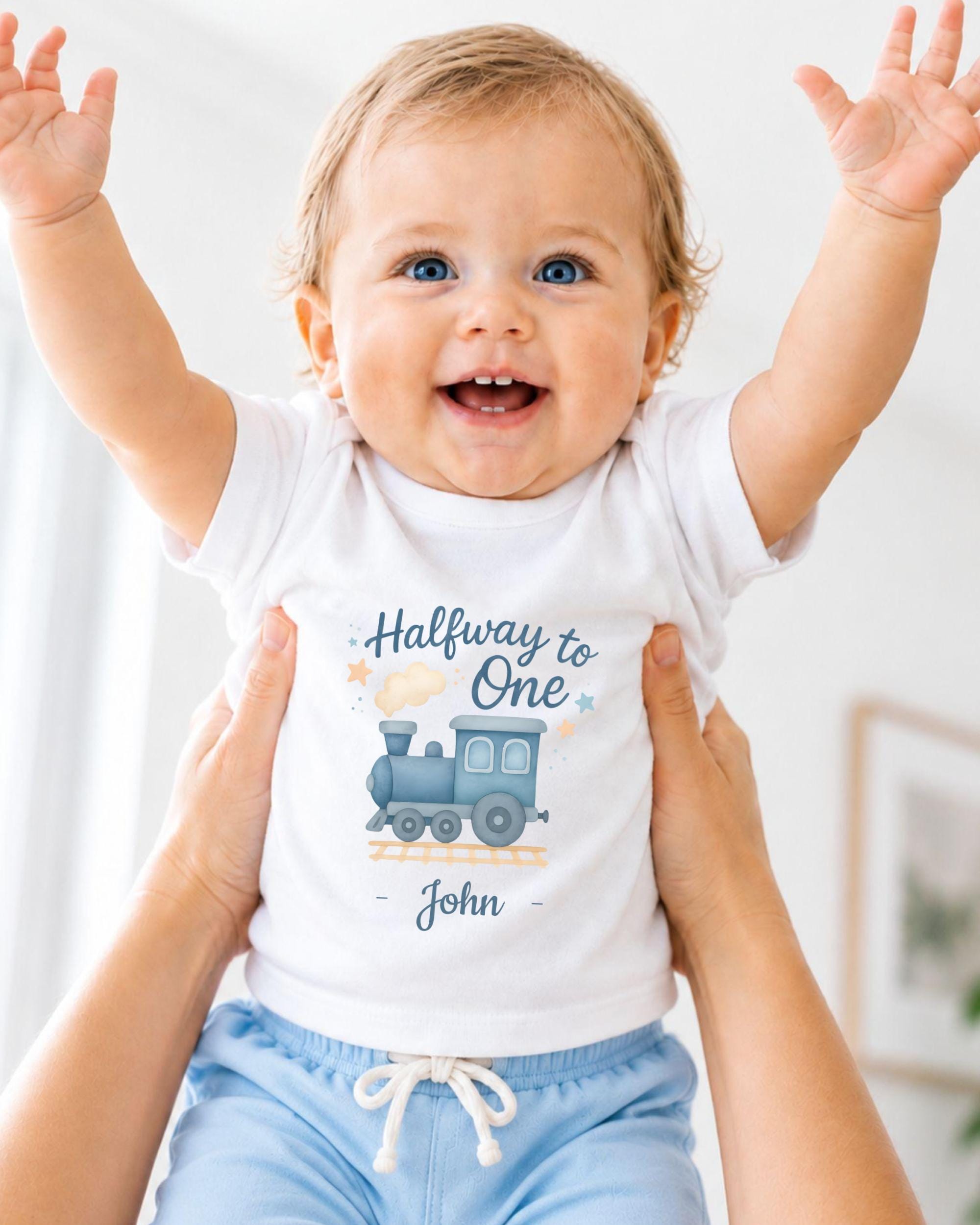 A smiling baby wearing a white t-shirt with a train graphic and the text "Halfway to One John" is being held up by someone's arms.