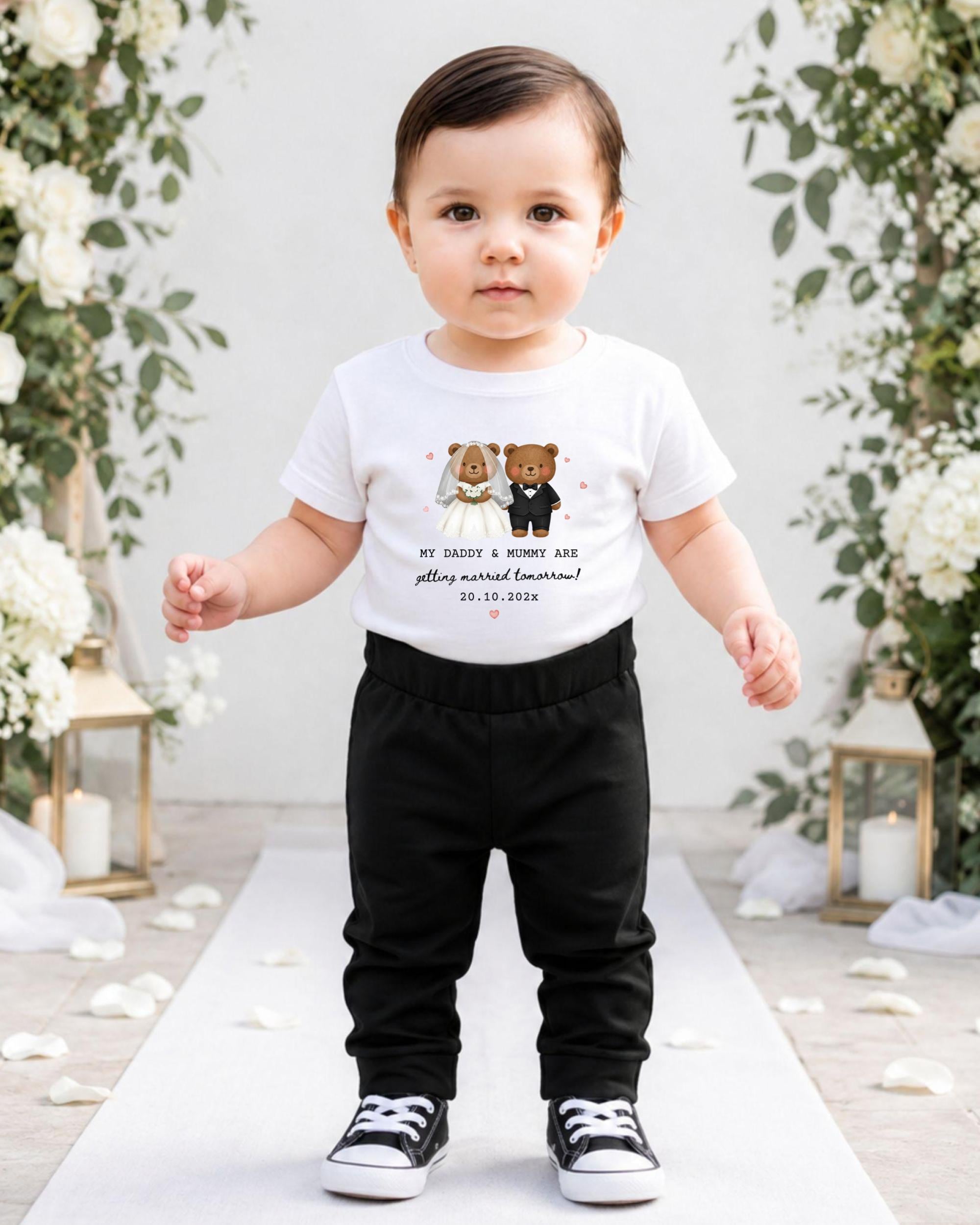 A young child wearing a white t-shirt with a graphic of two bears and text, standing on a white carpet in front of a floral backdrop.