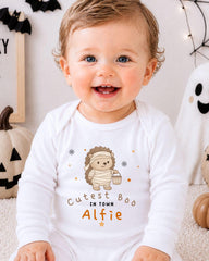 A smiling baby boy wearing a white onesie with a cartoon hedgehog and the text "Cute Bo in Town Alfie" is sitting in front of Halloween decorations.