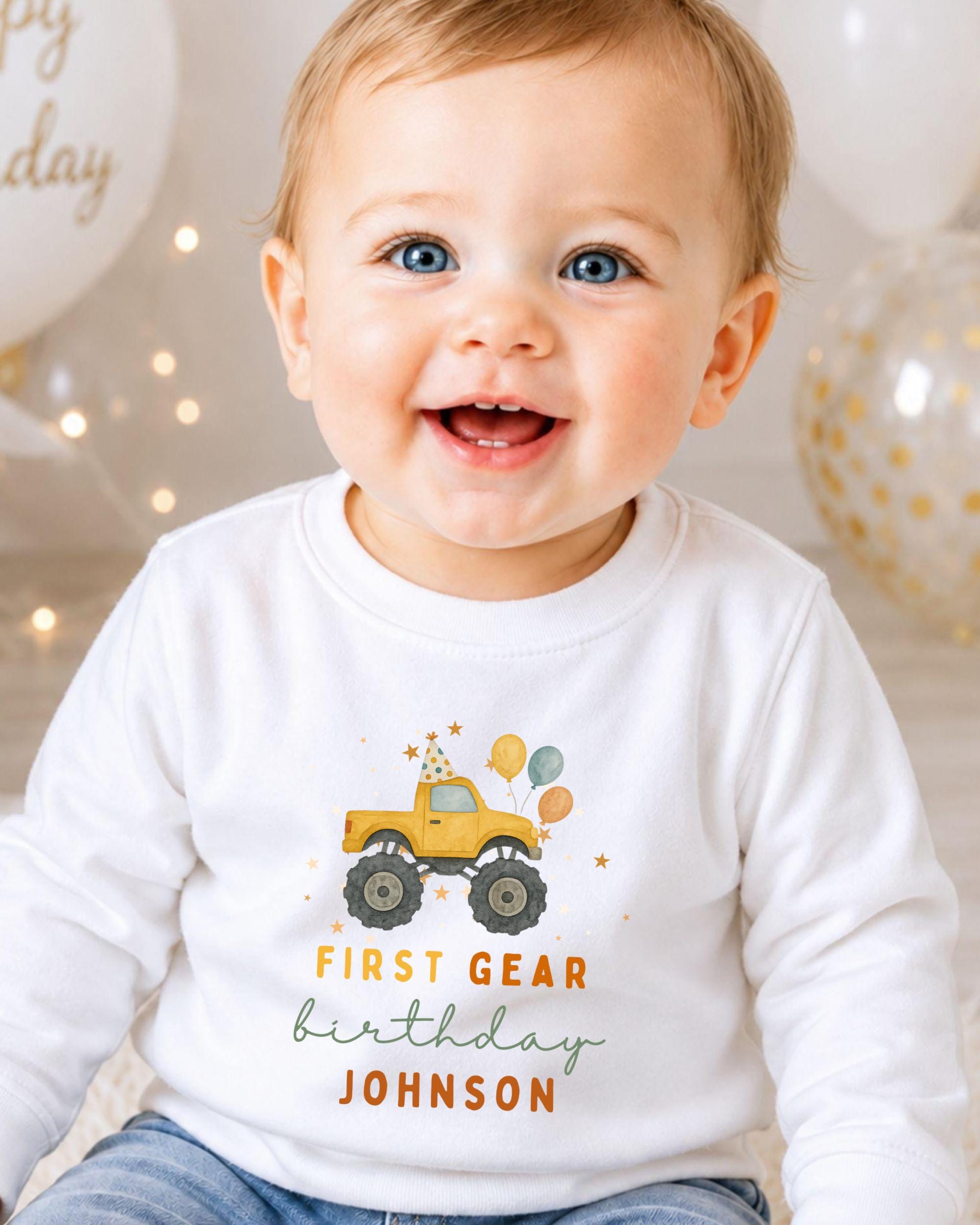 A smiling baby boy wearing a white shirt with a yellow monster truck graphic and the text "FIRST GEAR BIRTHDAY JOHNSON" printed on it.