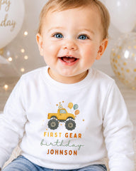 A smiling baby boy wearing a white shirt with a yellow monster truck graphic and the text "FIRST GEAR BIRTHDAY JOHNSON" printed on it.