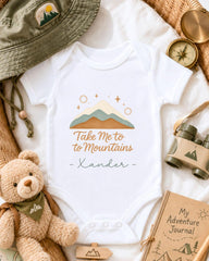 a baby onesie with a mountain and star design, surrounded by various outdoor-themed items such as a hat, compass, binoculars, and a teddy bear.