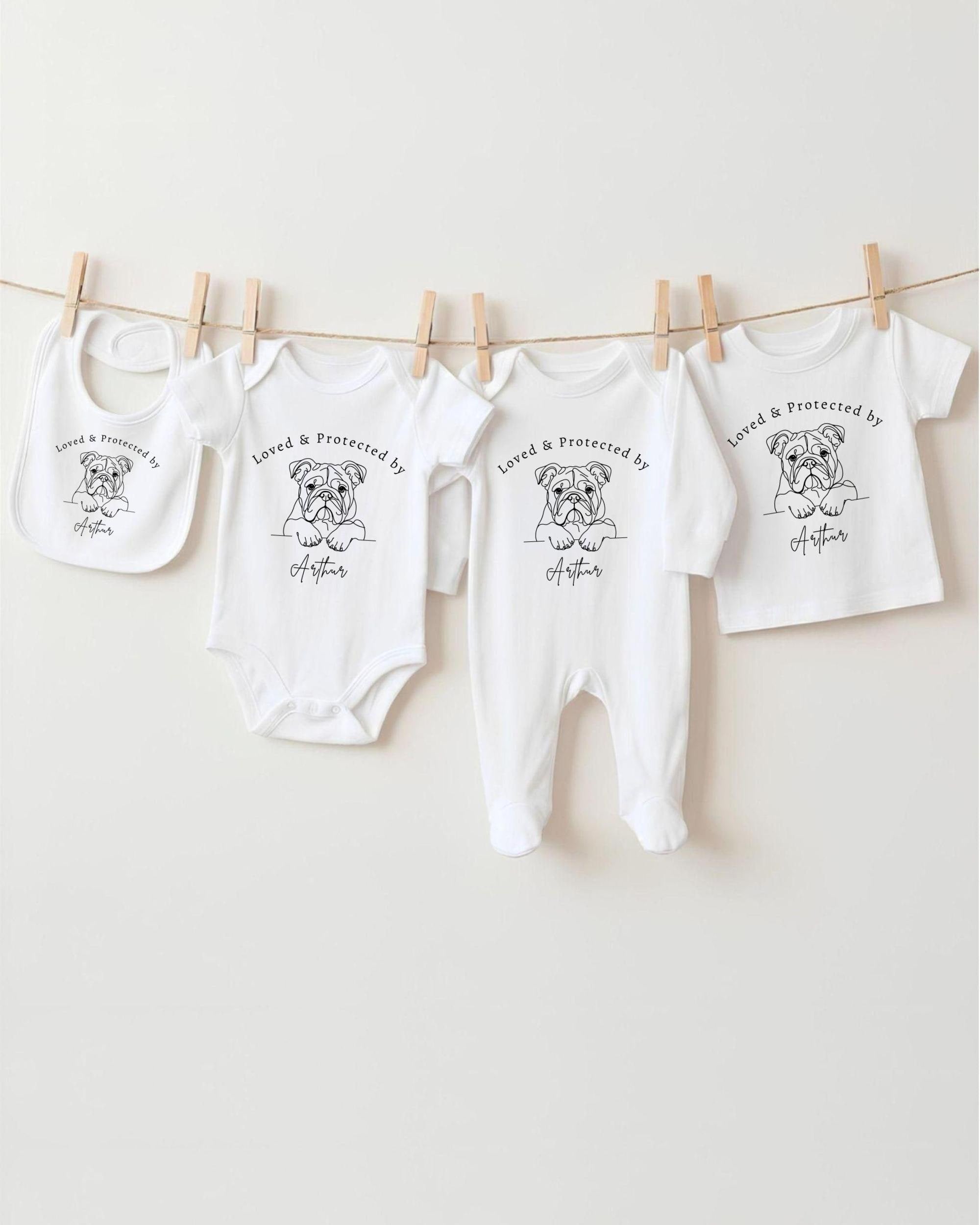 A collection of baby clothes and bibs hanging on a clothesline, featuring a dog illustration and the text "Loved & Protected by" printed on them.