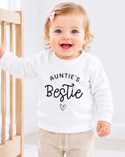 A smiling baby girl wearing a white sweatshirt with the text "Auntie's Bestie" printed on it.