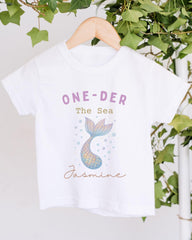 A white t-shirt with a mermaid tail design and the text "One-DER The Sea" printed on it, hanging on a wooden hanger.