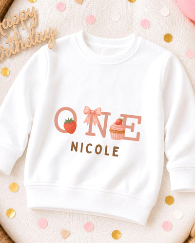 A white sweatshirt with a personalized text and graphics design, including a strawberry, cupcake, and the text "ONE NICOLE".