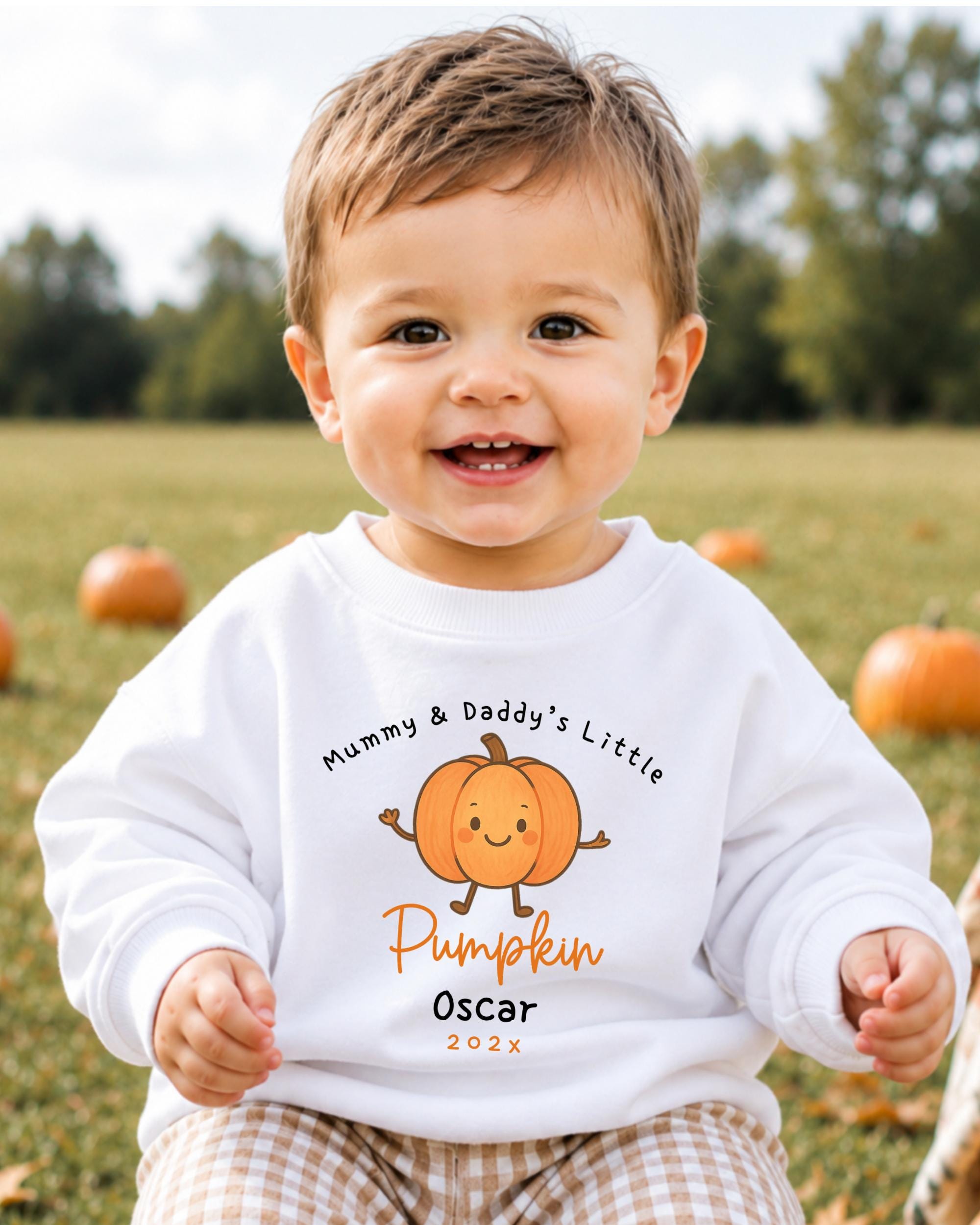 A smiling baby boy wearing a white sweatshirt with a pumpkin graphic and text.