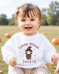 A smiling baby girl wearing a white sweater with a cartoon witch and the name " Jessie" printed on it, sitting in a field with pumpkins in the background.