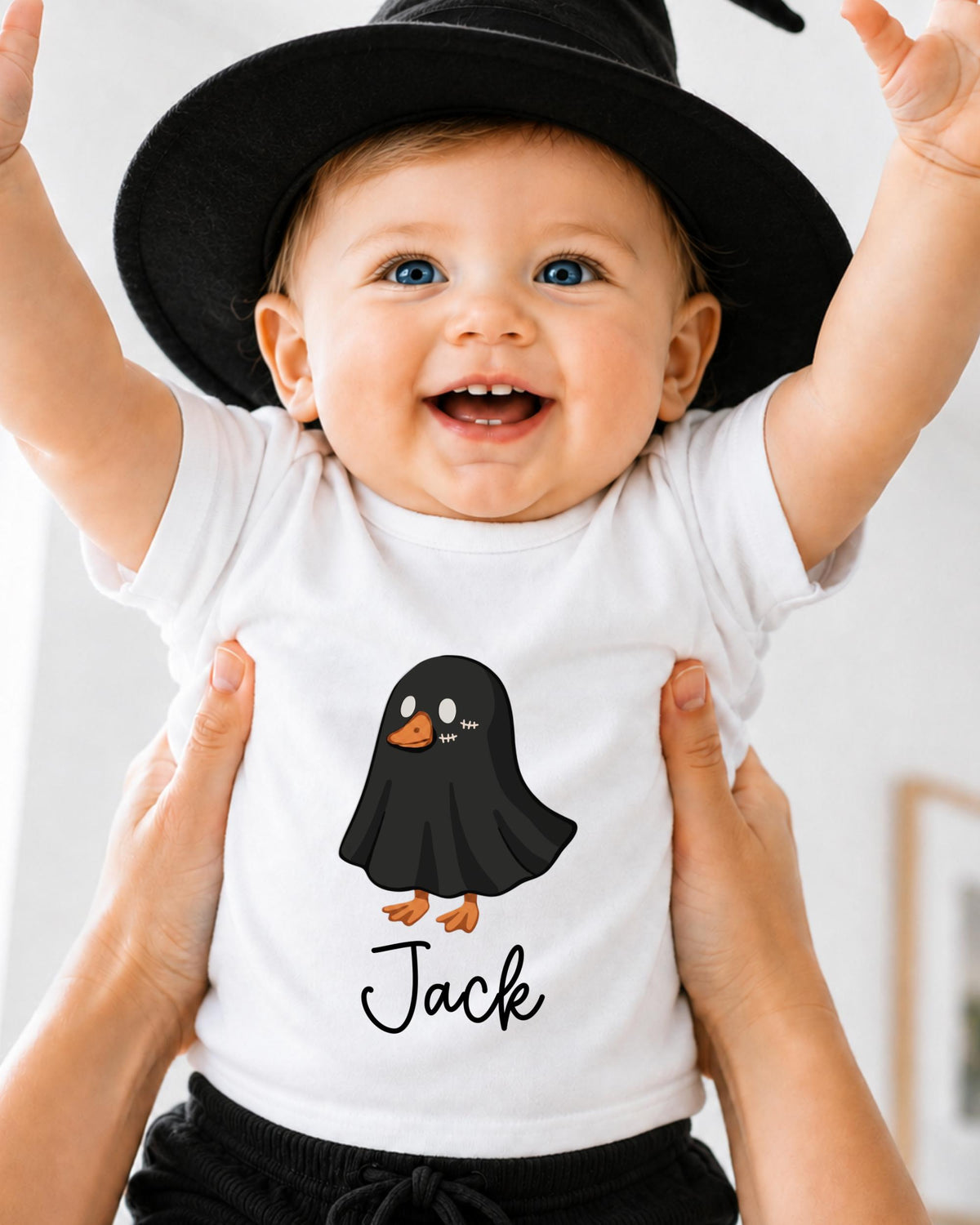 A smiling baby wearing a white t-shirt with a cartoon ghost and the name "Jack" printed on it, being held up by someone's hands.