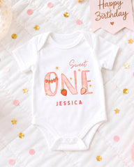 A white onesie with the text "Sweet One" and " Jessica" printed on it, along with a pink banner that says "Happy Birthday". The onesie is placed on a bed with confetti and stars scattered around it.