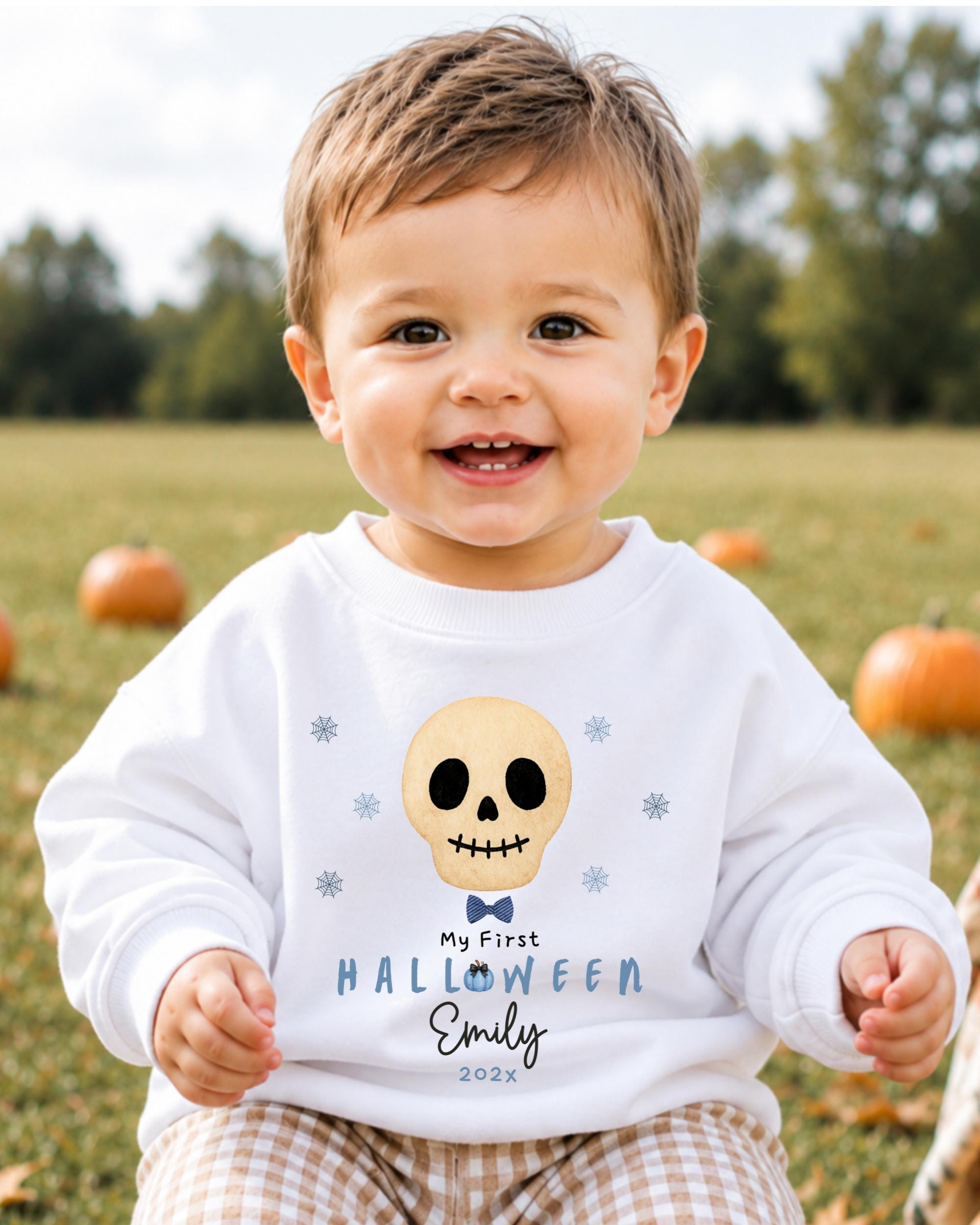 A smiling baby boy wearing a white sweatshirt with a skull design and text, sitting in a field with pumpkins in the background.