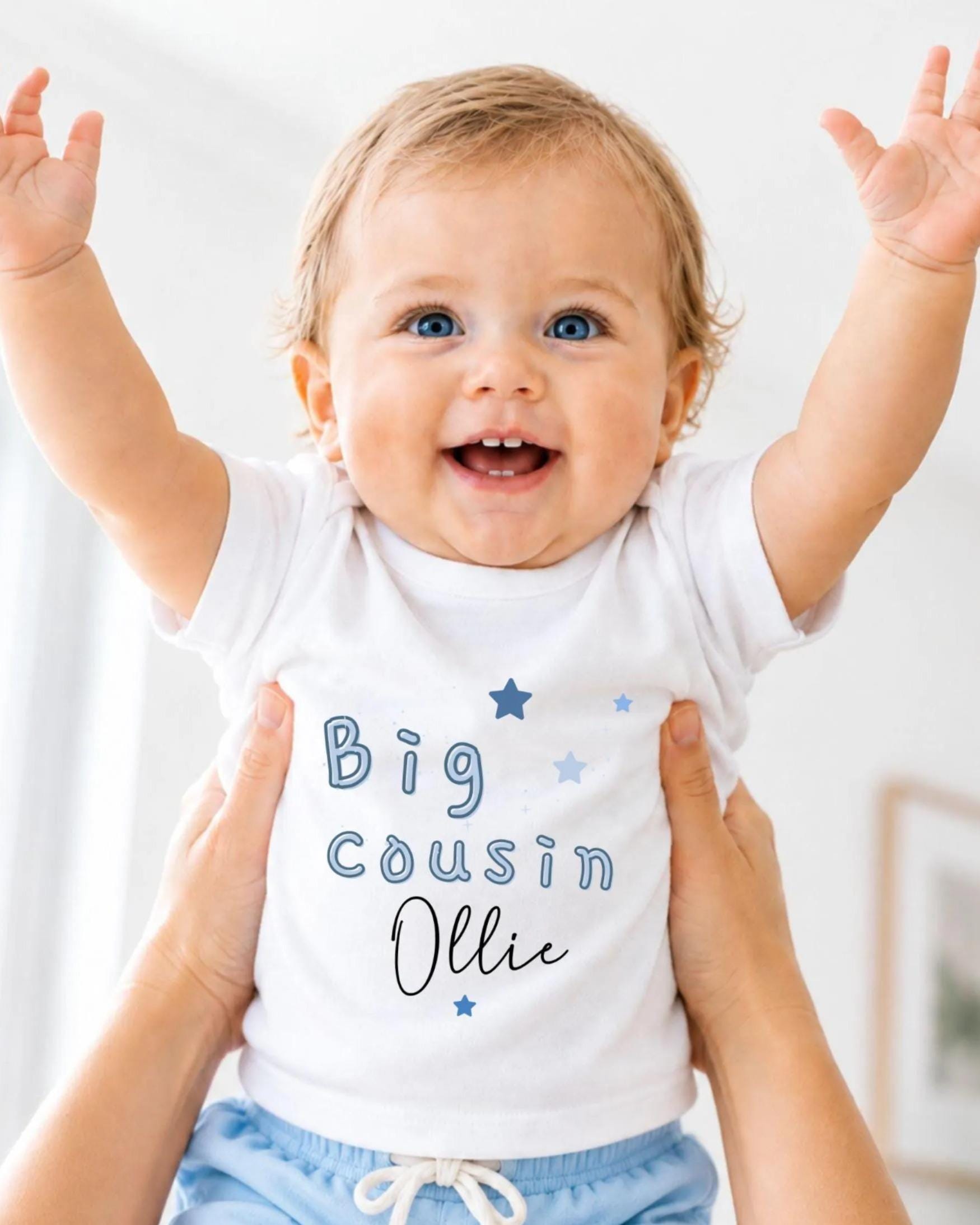 A smiling baby wearing a white shirt with the text "Big Cousin Ollie" is being held up by someone's arms.