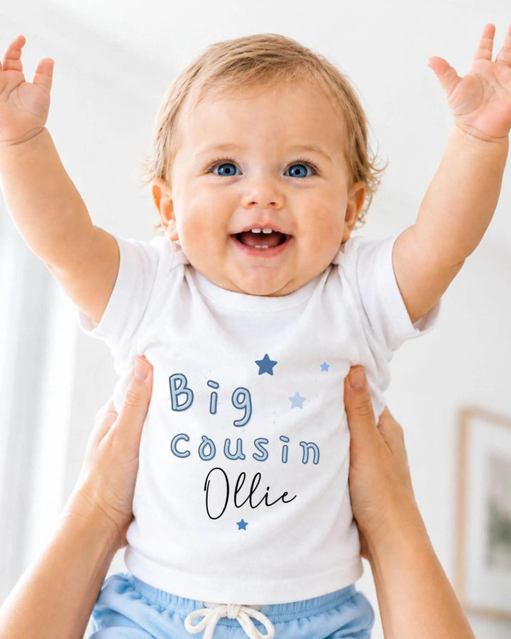 A smiling baby wearing a white shirt with the text "Big Cousin Ollie" is being held up by someone's arms.