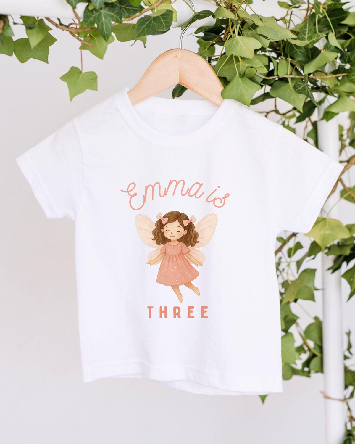 A white t-shirt with a graphic of a fairy and the text "Emma is THREE" printed on it, hanging on a wooden hanger.