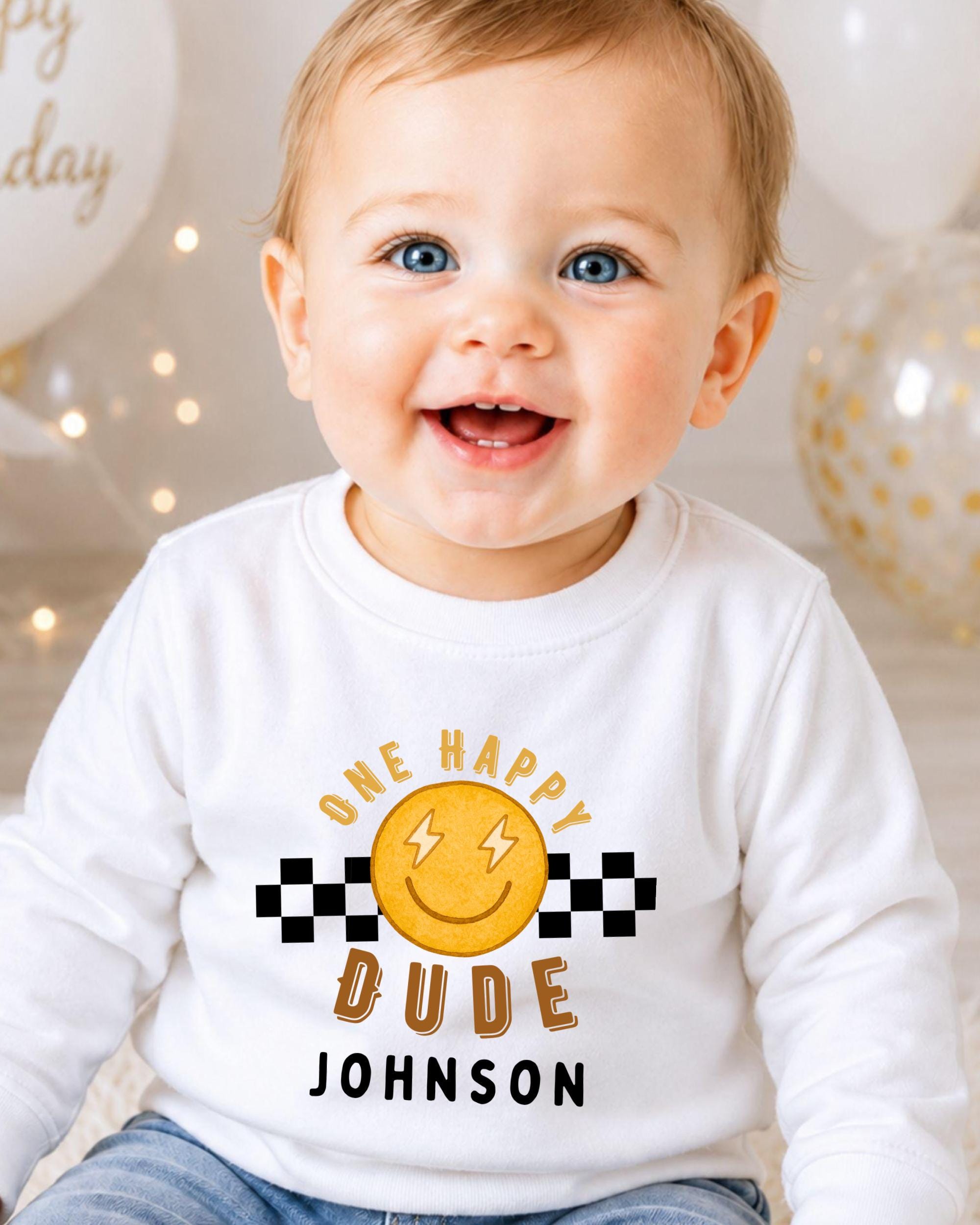 A smiling baby boy wearing a white shirt with a happy face and text that says "One happy dude Johnson".