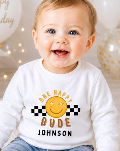 A smiling baby boy wearing a white shirt with a happy face and text that says "One happy dude Johnson".
