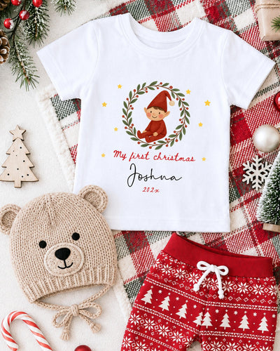 A white t-shirt with a Christmas-themed design, a beige knit hat with a Christmas tree, and red and white patterned shorts, all arranged on a plaid blanket with Christmas decorations.