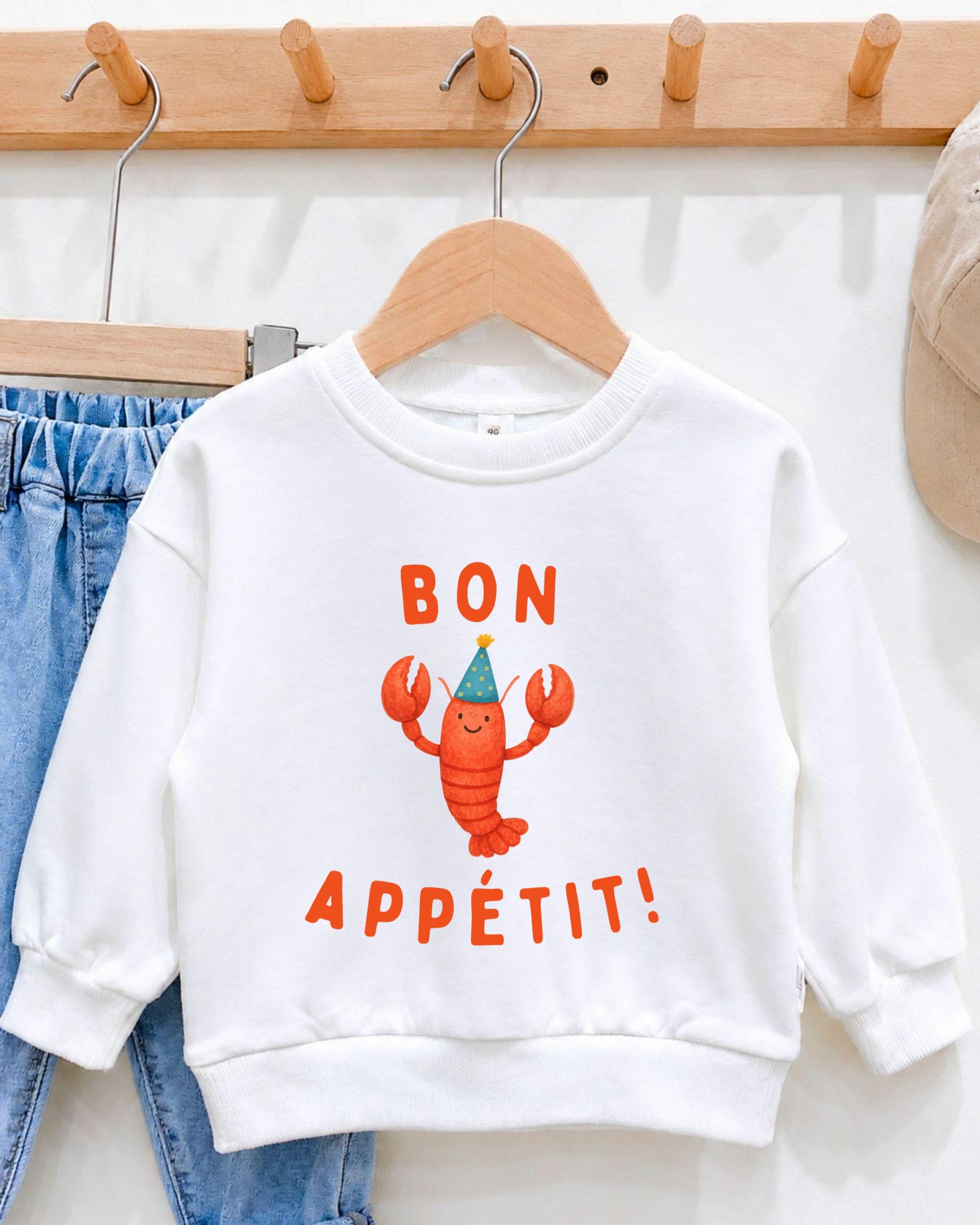 A white sweater with a cartoon lobster and the text " bon appetit!" printed on it, hanging on a wooden hanger.