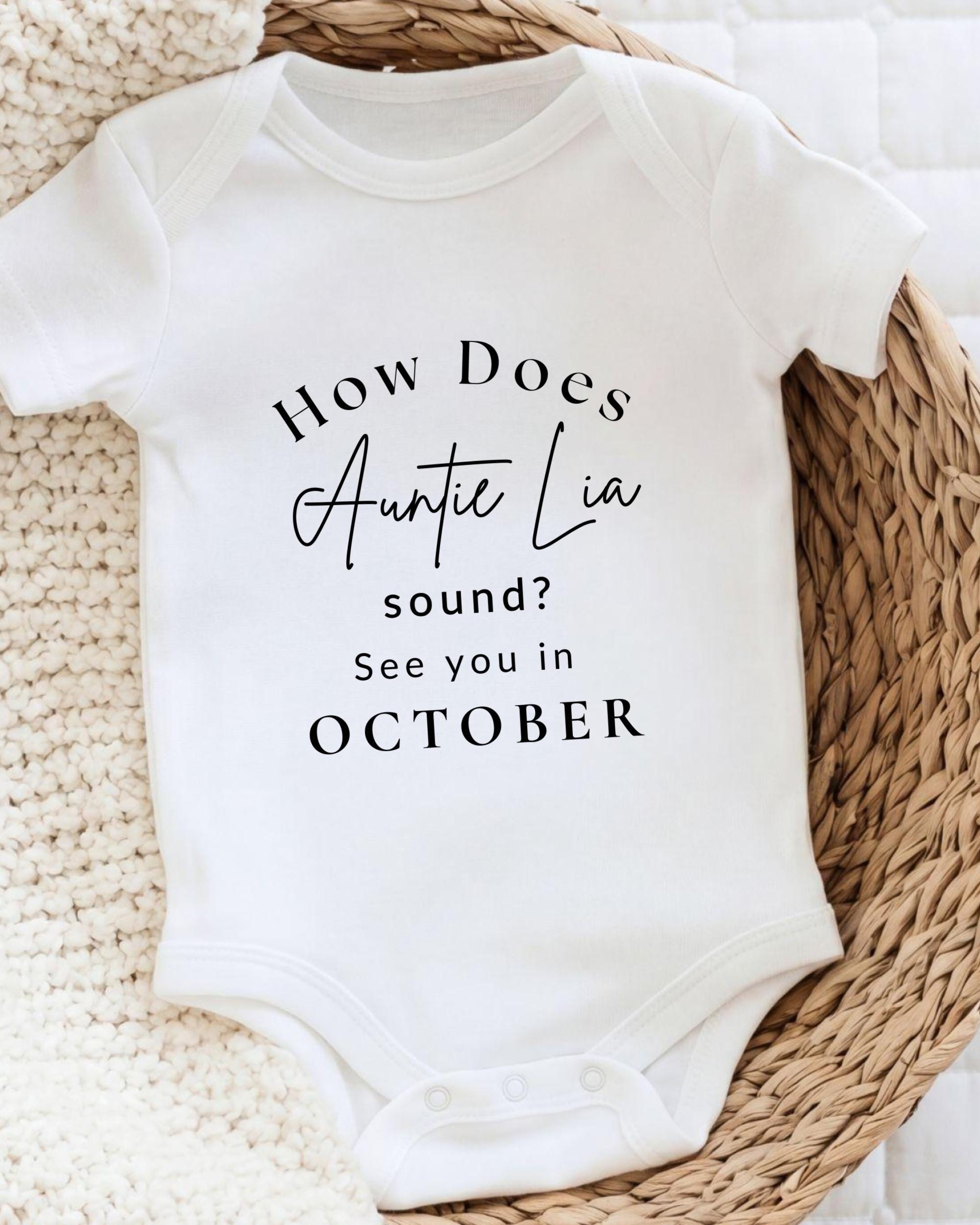 A white baby onesie with text printed on it, placed on a woven basket.