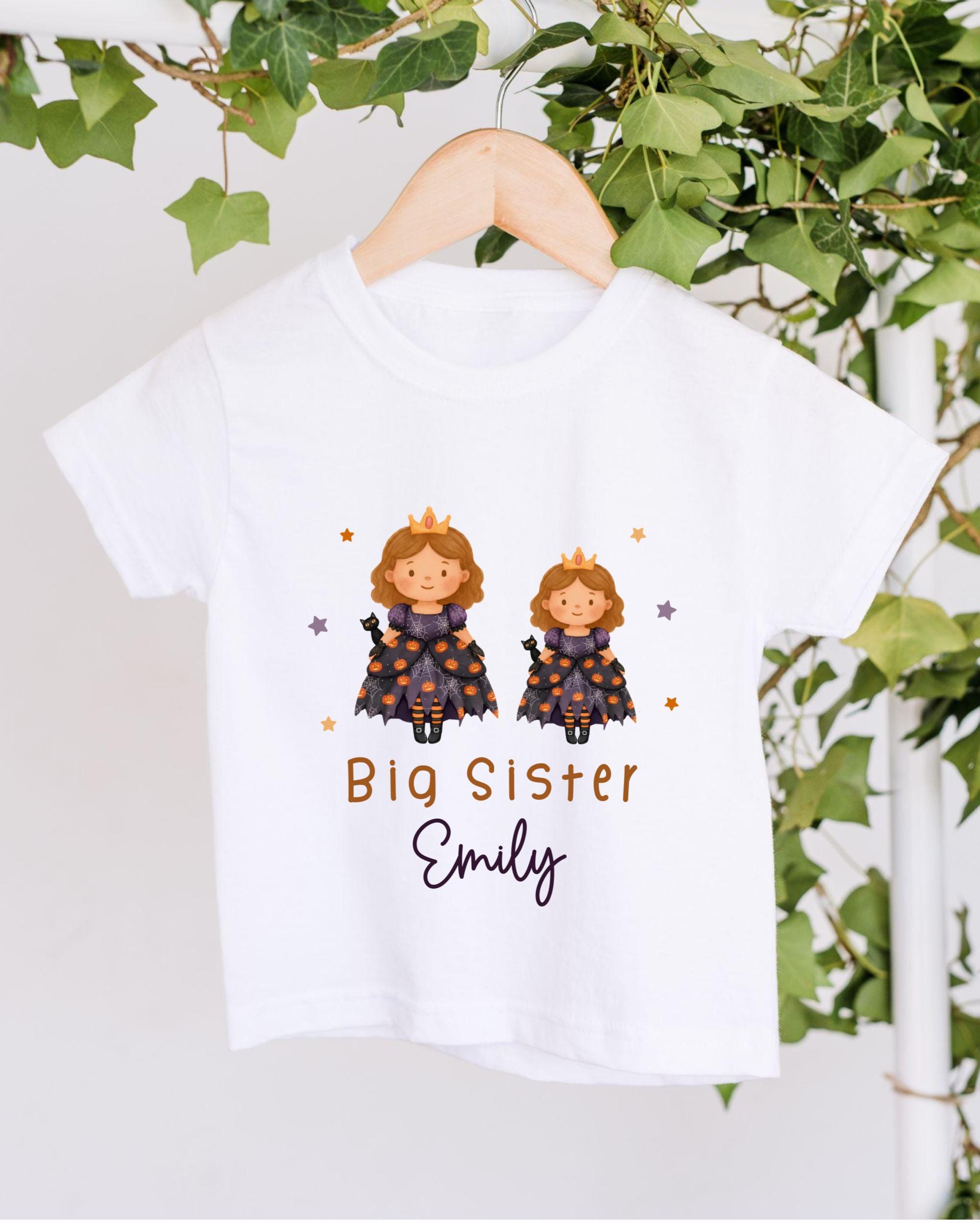 A white t-shirt with a graphic design featuring two cartoon princesses wearing crowns, surrounded by stars and the text "Big Sister Emily".