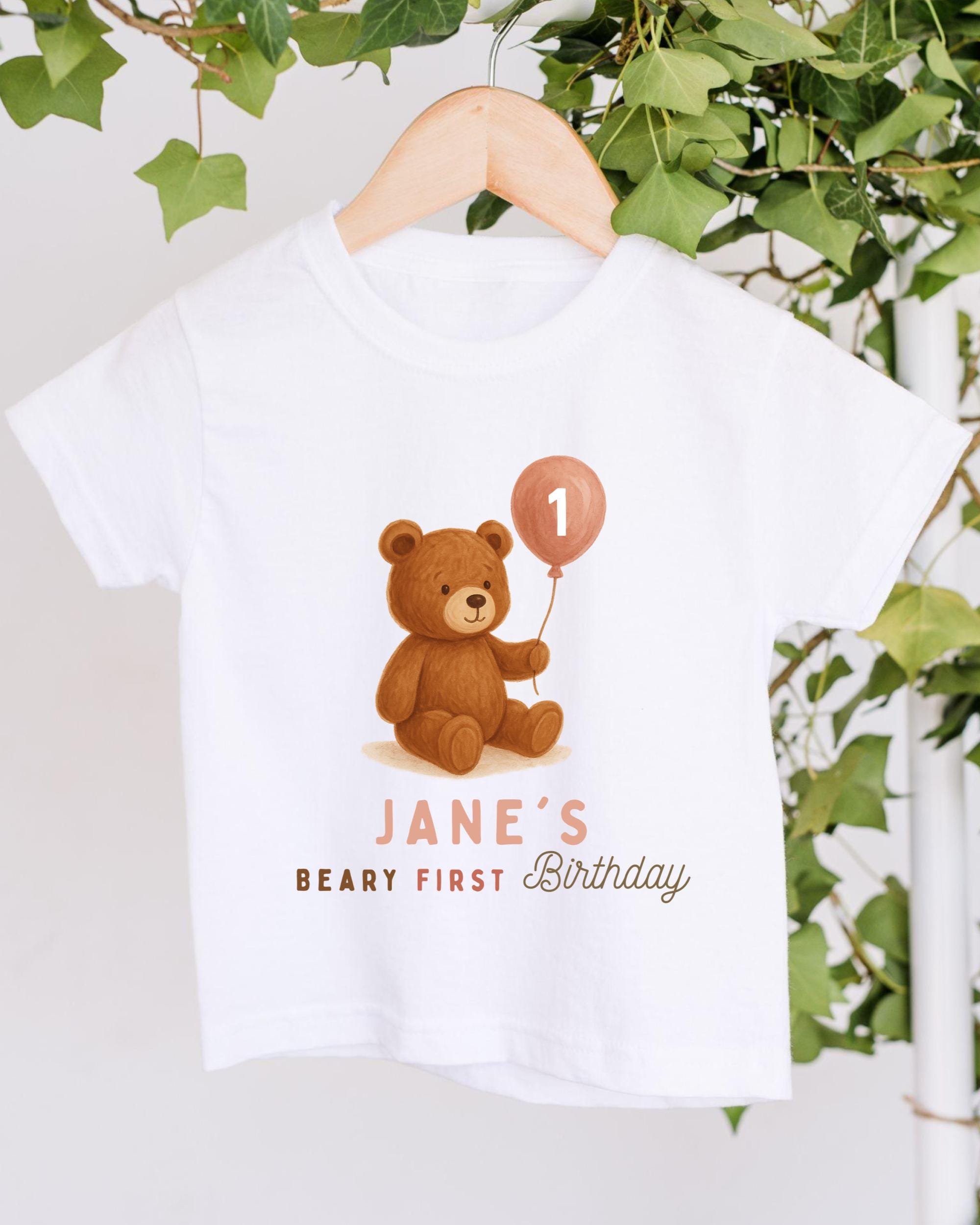 A white t-shirt with a teddy bear and a birthday balloon printed on it, along with the text "JANE'S BEARY FIRST BIRTHDAY".