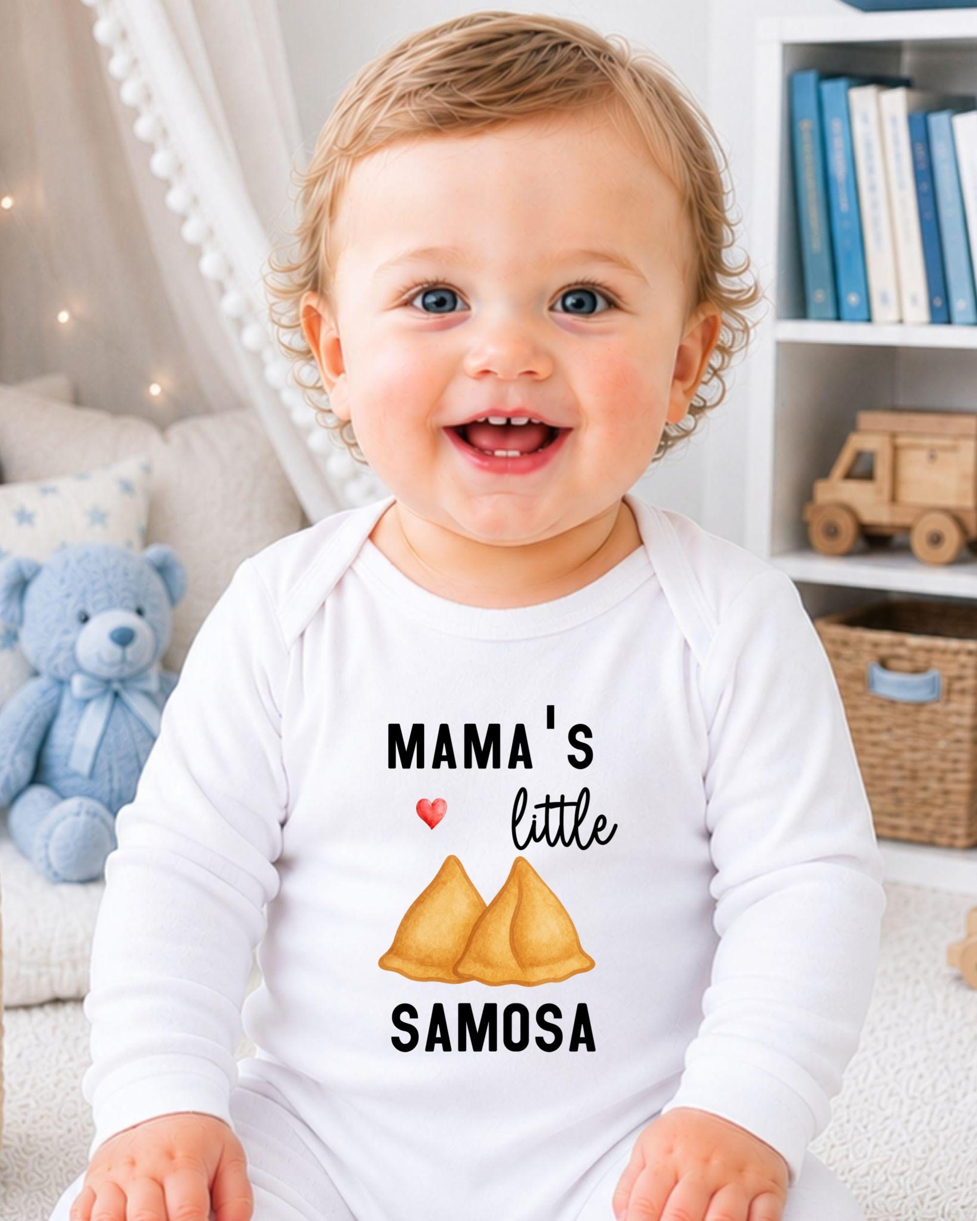 A smiling baby wearing a white onesie with a printed design of samosas and the text "Mama's little samosa" is sitting in a room with a teddy bear and bookshelf in the background.