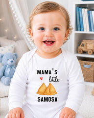 A smiling baby wearing a white onesie with a printed design of samosas and the text "Mama's little samosa" is sitting in a room with a teddy bear and bookshelf in the background.