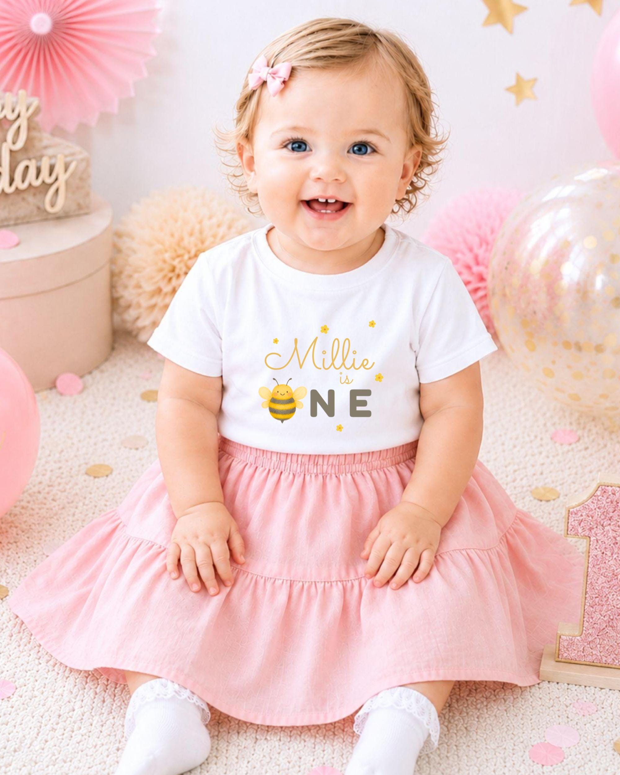 A smiling baby girl wearing a white t-shirt with a bee design and a pink skirt, sitting on a pink and white background with balloons and confetti.