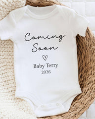 A white baby onesie with the text "Coming Soon Baby Terry 2026" printed on it, placed on a woven basket.