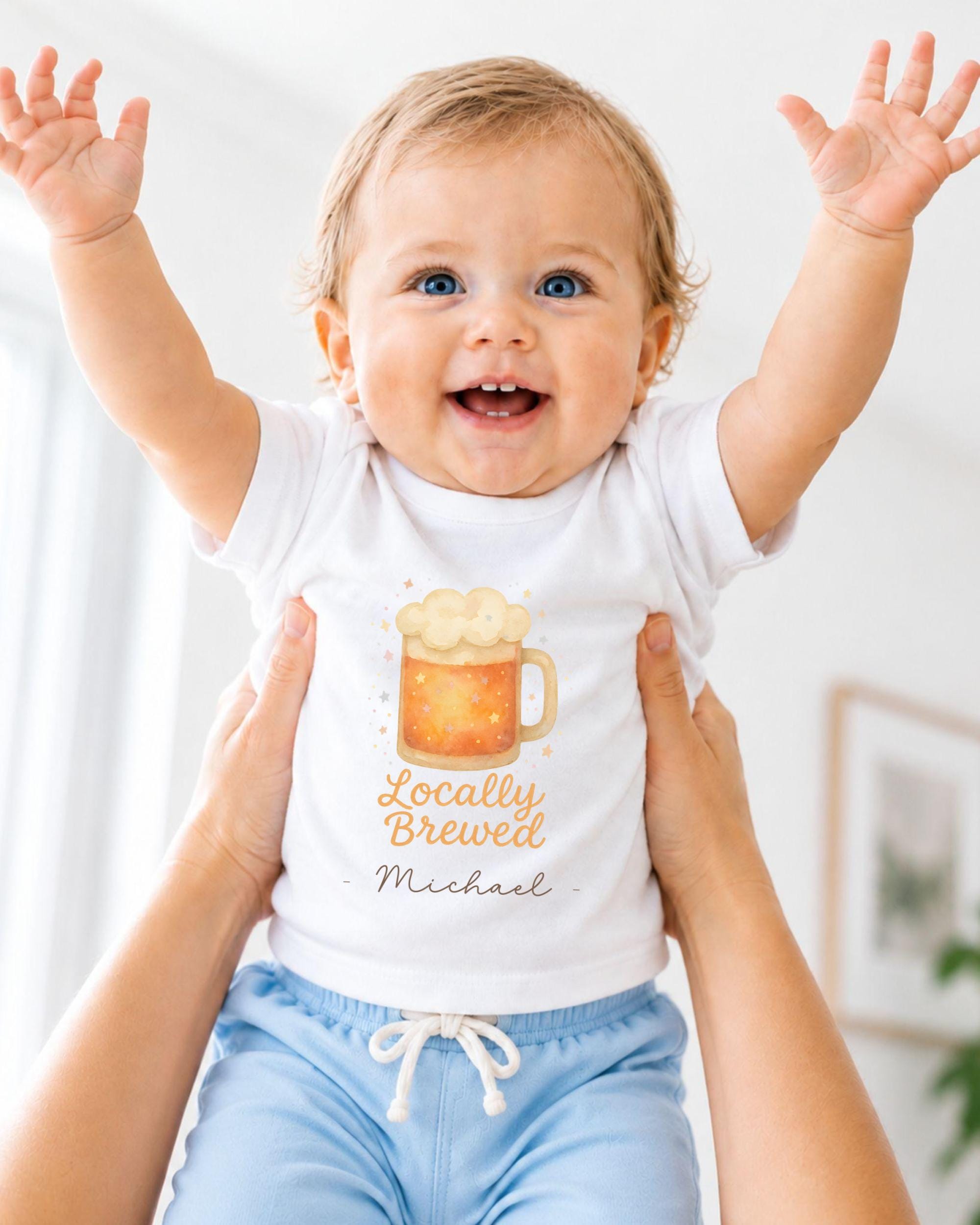 A smiling baby wearing a white shirt with a beer mug graphic and the text "Locally Brewed Michael" is being held up by someone's arms.