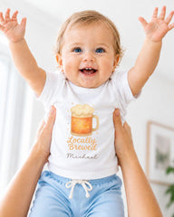 A smiling baby wearing a white shirt with a beer mug graphic and the text "Locally Brewed Michael" is being held up by someone's arms.
