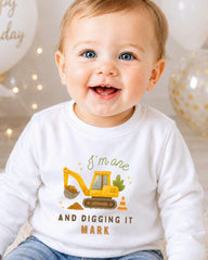 A smiling baby boy wearing a white shirt with a construction vehicle graphic and text.